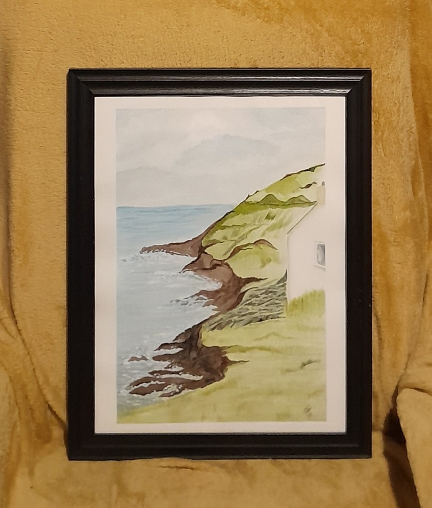 Framed watercolour painting of a coastal cliff with green grass and a white cottage